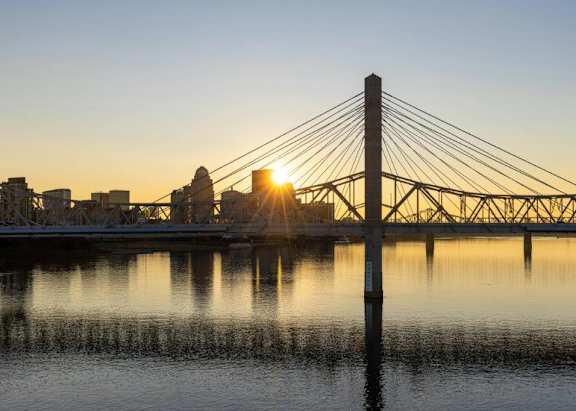 A bridge spans over calm waters with the sun setting in the background.