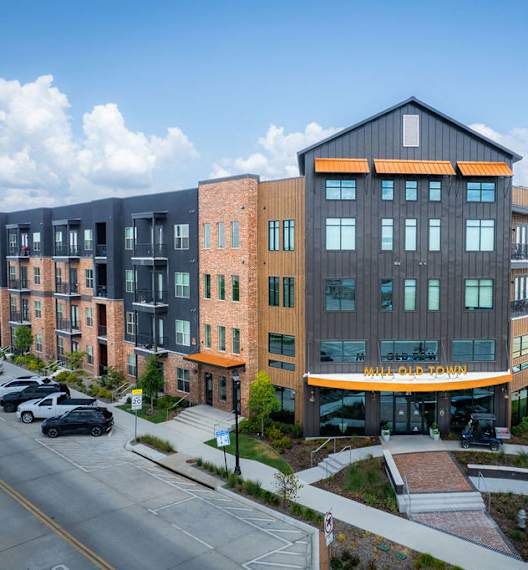 A modern multi-story building with a mix of brick and panel construction is situated on a street corner with cars parked in front.