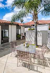 A patio with a table and chairs is surrounded by a house and a palm tree.