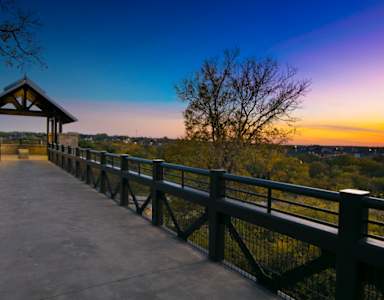 A wooden gazebo sits on a bridge with a view of the city in the distance.