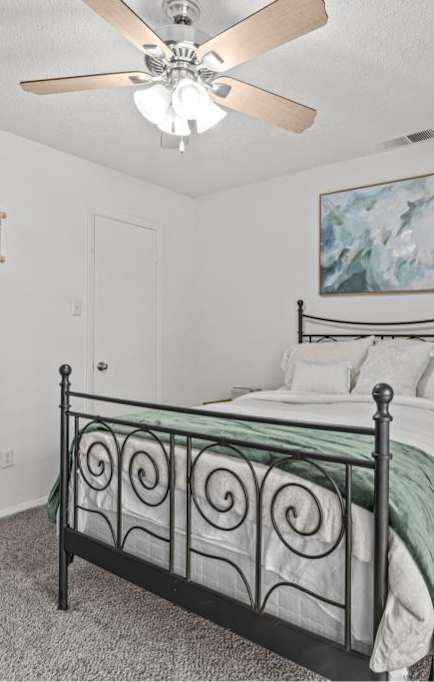 A bedroom with a bed, a ceiling fan, and a painting on the wall.