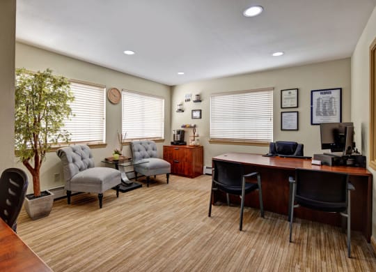 Office here at Greenbriar Apartments with a wooden desk, black chairs, and a computer. Two gray armchairs and a potted tree add warmth. Certificates and a clock adorn the walls.