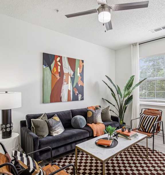 A living room with a black couch, a painting on the wall, a ceiling fan, and a large window with a plant in front of it.