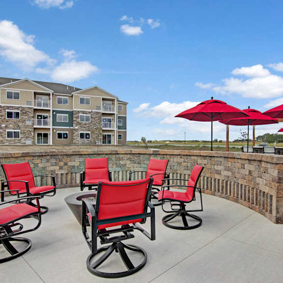 A patio with a firepit with red chairs surrounding it, and dining tables and chairs with red umbrellas with the apartment buildings in the background at the Club at Heritage Apartment Homes.