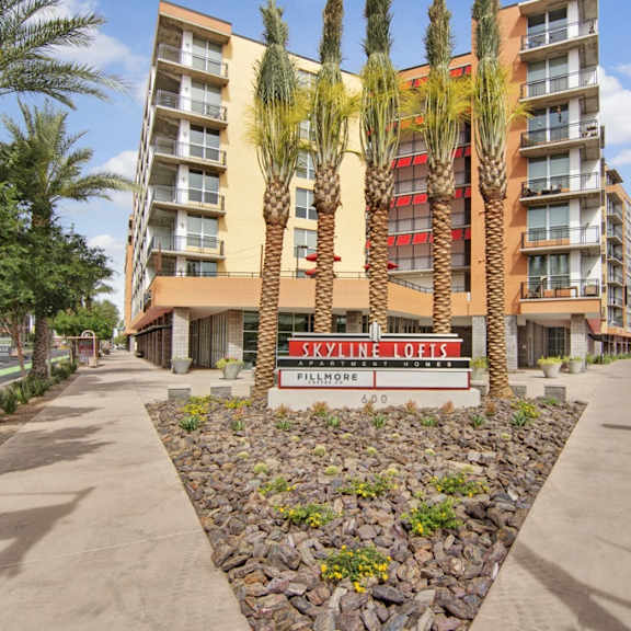 The front view of Skyline Lofts with a view of the apartments, the front sign, and palm trees behind a triangle of rocks that houses plants growing in them. 