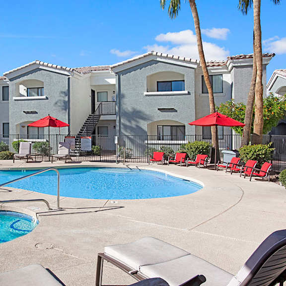 The bright blue pool and hot tub surrounded by red chairs and umbrellas with palm trees at Pala Mesa Apartments. 