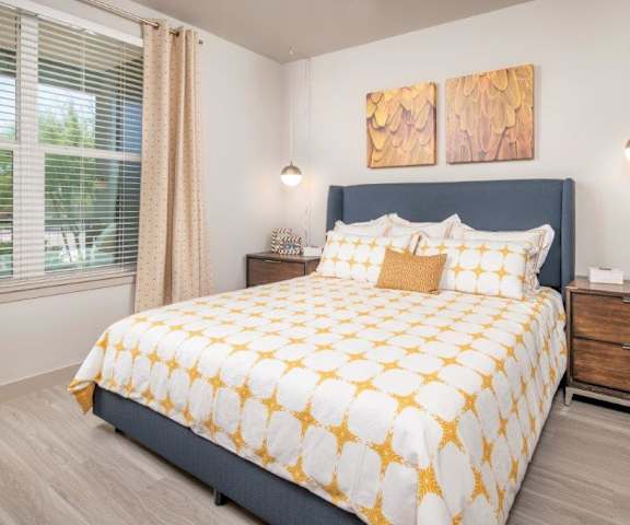 A spacious bedroom with a queen-size bed with a gold star and white bedspread, a dresser, wall art, lamps, decorations, window with yellow curtains, and nightstands with lamps at the Flats at San Tan Apartment Homes. 