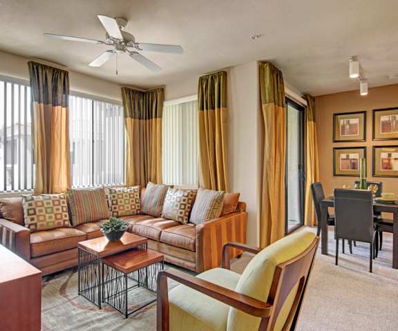 The living room with an orange color scheme, a couch, plush chairs, coffee table, decorations, a TV, and view of the dining room at Natura Villas Apartments. 
