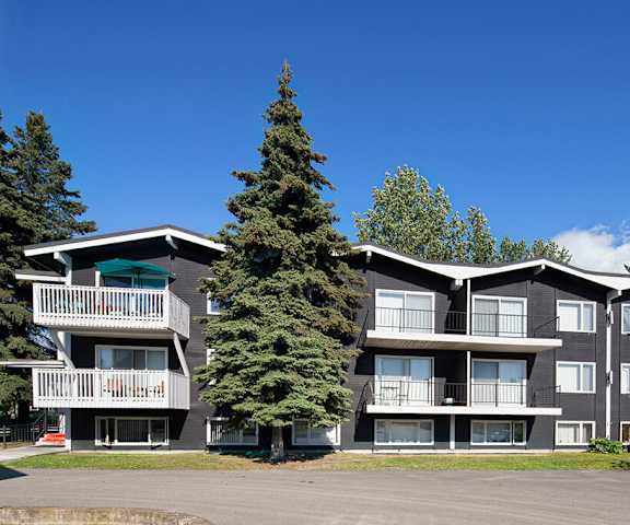 The black modern apartment buildings surrounded by the trees with grass and a picnic table in the front at the Alaskan Apartment Homes. 