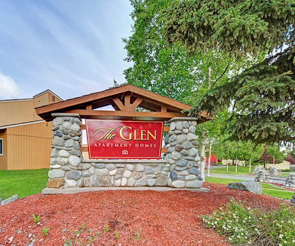The exterior sign with trees and surrounded by greenery, trees, and flowers with the buildings in the background of the Glen Apartment Homes. 