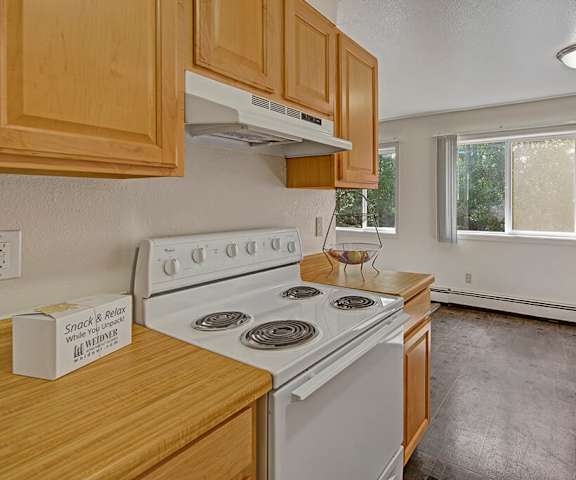 A view of the empty carpeted living room with a window and the kitchen with white appliances and wooden large cabinets at the Glen Apartment Homes. 
