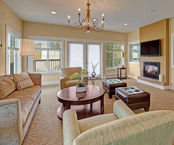 The amazing resident lounge with a fireplace, leather footrests, couches, decorations, TV, a chandelier, yellow walls, a glass door, large windows with curtains, plush chairs, and coffee tables at the Glen Apartment Homes. 