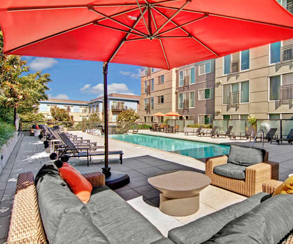 The side view of the turquoise outdoor pool with pool chairs, tables, and chairs with the apartments in the background at Leilani Apartment Homes. 
