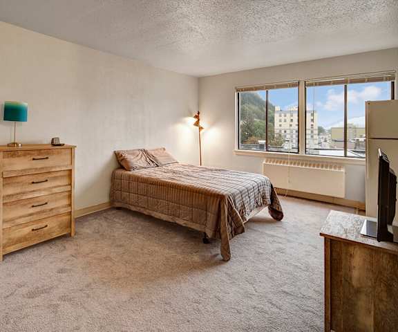 A bedroom with a twin-size bed, a dresser, decorations, window, and nightstands at Mendenhall Tower Apartments. 