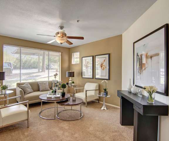 The living room with a couch, plush chairs, coffee table, decorations, a TV, and view of the parking lot from the windows at Mirador Apartments. 