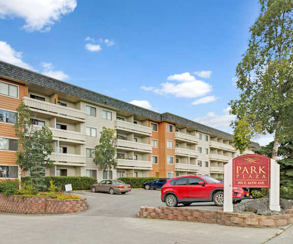 The exterior sign with trees and surrounded by greenery with the buildings and parking lot in the background of Park Plaza Apartments. 