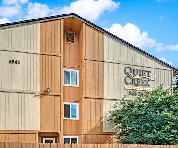 The exterior of the Quiet Creek Apartment Homes showing the sign for the apartments on the side of the building surrounded by trees. 