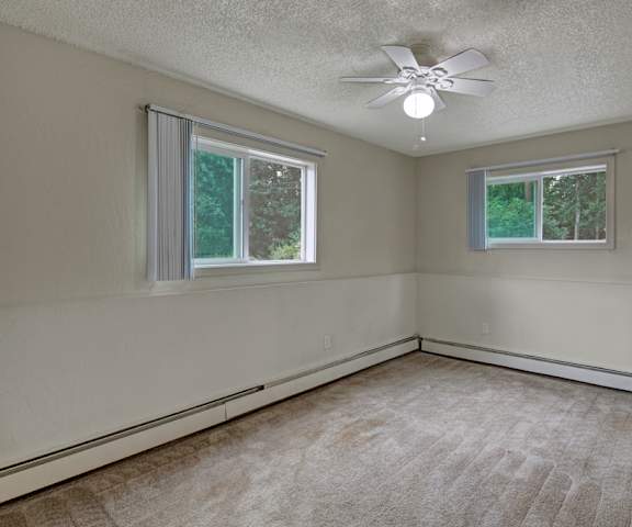 An empty carpeted bedroom with two windows and a ceiling fan at Rancho Tudor Apartments. 