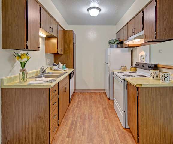 A large kitchen with dishes, candles, flowers, white appliances, a light counter with lots of room, and spacious wooden cabinets at Rancho Tudor Apartments. 