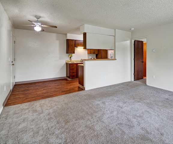 A view of the empty carpeted living room with a window and the kitchen with black appliances and wooden large cabinets at Rancho Tudor Apartment Homes. 