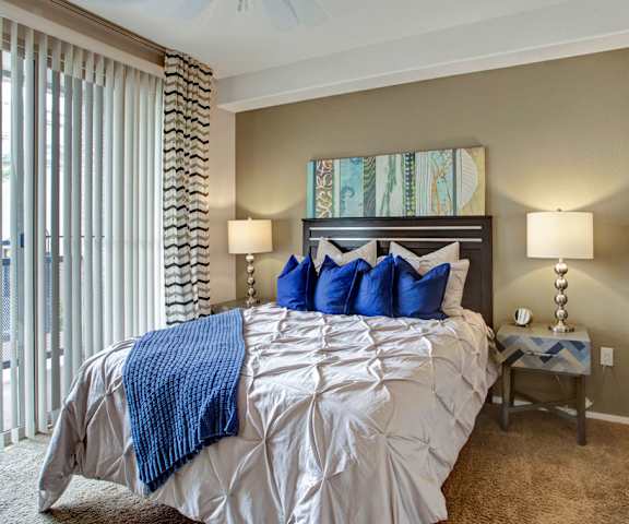 A spacious bedroom with a white and blue queen-size bed, a dresser, wall art, lamps, decorations, window, and nightstands at Rio Paradiso Apartment Homes. 