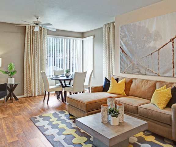 The spacious living room with a couch, windows with curtains, plush chairs, coffee table, wall art, overhead lighting with a ceiling fan, decorations, a TV, and view of the dining room with a table, chairs, and place settings for four, and the kitchen at Rio Paradiso Apartment Homes. 