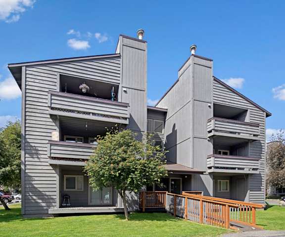 The exterior of the Russian Jack gray apartment homes surrounded by grass and trees with a pathway leading to the front door. 