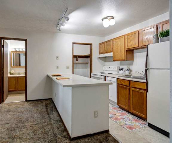A spacious cozy kitchen with white appliances, kitchen utensils, overhead lighting, light countertops with lots of room, a bar counter with two places set, and large wooden cabinets at Russian Jack Apartment Homes. 
