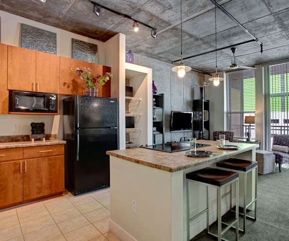 A spacious, industrial kitchen with black appliances, marble countertops with lots of room, a kitchen island with barstool, light wooden cabinets, decorations, and a view of the well-decorated living room at Skyline Lofts Apartment Homes. 