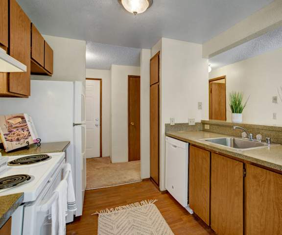 A cozy kitchen with white appliances, a white rug, kitchen utensils, overhead lighting, light countertops with lots of room, decorative plants, and large wooden cabinets at Soundview Apartment Homes. 