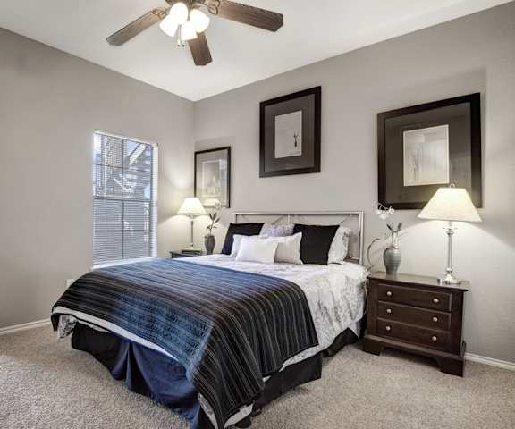 A spacious bedroom with a queen-size bed with a navy and white bedspread, a dresser, wall art, lamps, decorations, overhead lighting with a fan, window, and nightstands with lamps at Stoneleigh on May Apartment Homes. 