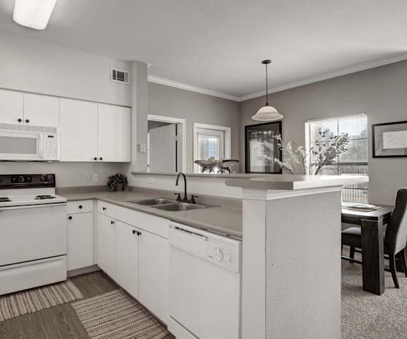 A spacious, modern, gray kitchen with white appliances, kitchen utensils, overhead lighting, light gray countertops with lots of room, a bar counter with barstools, and large, white wooden cabinets at Stoneleigh on May Apartment Homes. 