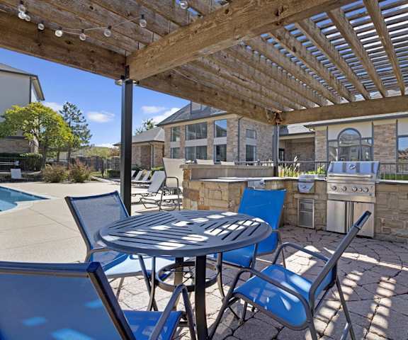 The grilling station underneath a lattice cover with a table and blue chairs with the pool area in the background at Stoneleigh on May Apartment Homes. 