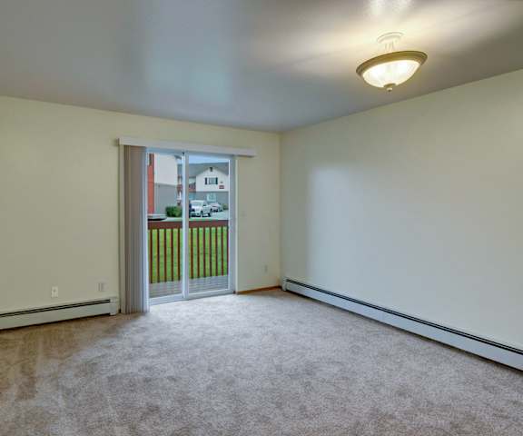 The empty carpeted living room with overhead lighting and a sliding glass door to the patio at Strawberry Lane Apartment Homes. 