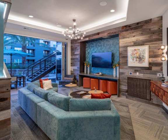 The resident lounge with artwork, a large mirror, interesting lighting, orange footstools, a coffee table, a TV, and a wraparound light blue couch with stairs in the background at the Flats at San Tan Apartments. 