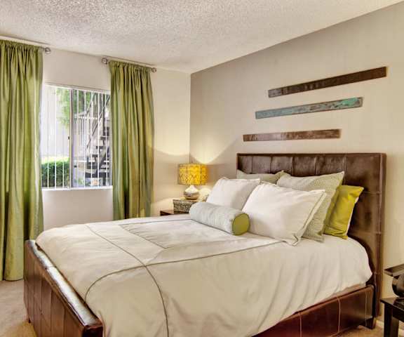 A bedroom with a queen-size bed, a dresser, decorations, window with satiny green curtains, and nightstands at Paradise Falls Apartments. 