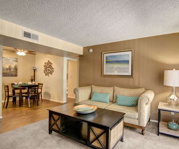 The living room with a couch, plush chairs, coffee table, decorations, a TV, and view of the dining room with a table and chairs and fully set places at Paradise Falls Apartments. 