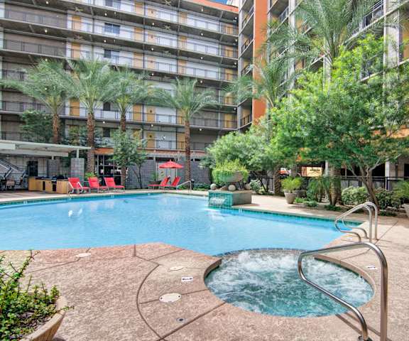 The large, turquoise outdoor pool surrounded by palm trees, red pool chairs, tables, red umbrellas, a small hot tub, with the apartments in the background at Skyline Lofts Apartment Homes. 