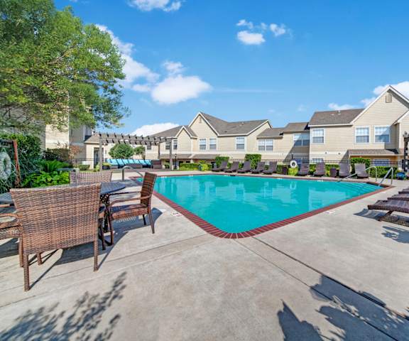The large, turquoise outdoor pool surrounded by trees, dark pool chairs, tables and chairs with the apartments in the background at Summit Pointe Apartment Homes. 