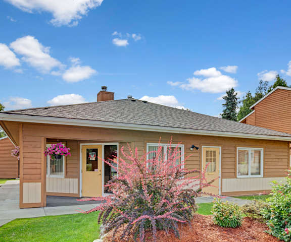 The exterior of the orange leasing office with hanging flowers surrounded by grass and plants and blue sky above at Wildwood Estates Apartments. 