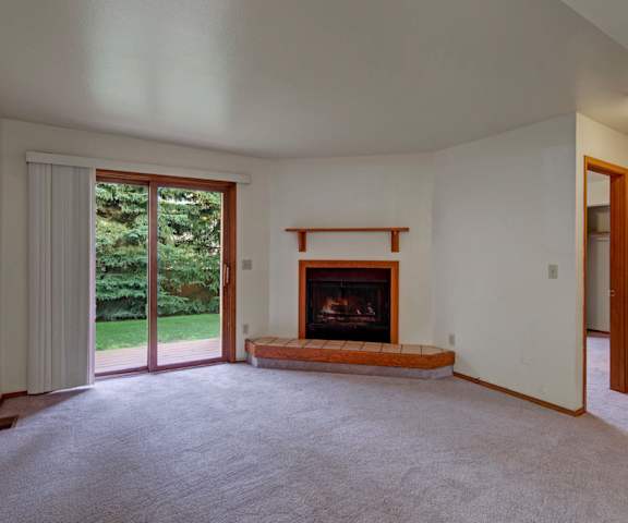An empty living room with carpeting, fireplace, overhead lighting, a door to the patio at Wildwood Estates Apartment Homes. 