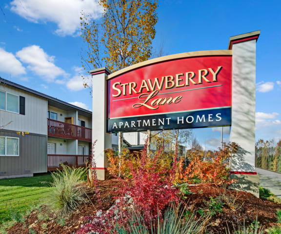 The exterior, red sign with trees and surrounded by greenery and flowers with the buildings in the background of Strawberry Lane Apartment Homes. 