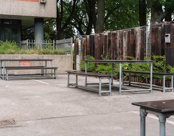 A concrete bench with a metal frame is in the foreground of a concrete area with a fence and trees in the background.