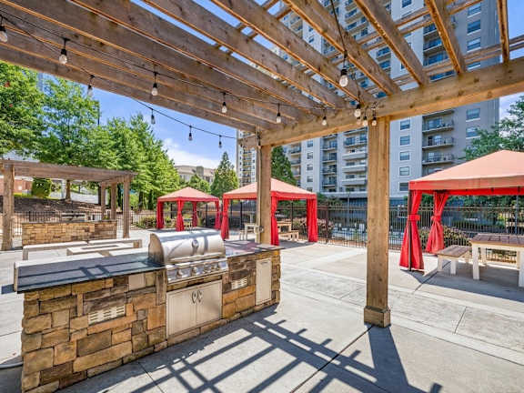 A wooden pergola with red awnings and string lights is over a stone barbecue area.