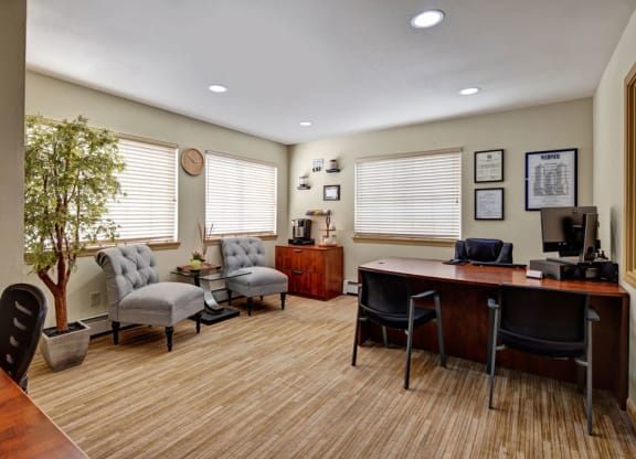 Office here at Greenbriar Apartments with a wooden desk, black chairs, and a computer. Two gray armchairs and a potted tree add warmth. Certificates and a clock adorn the walls.