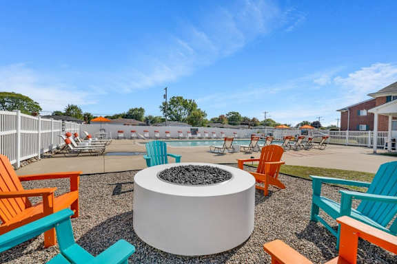 A white fire pit sits in the middle of a gravel area with orange and blue chairs surrounding it.