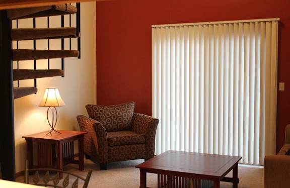  A living room with a chair, spiral staircase, and vertical blinds covering patio door