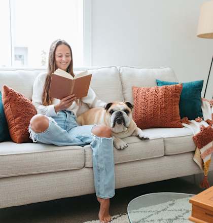 A woman is reading a book on a couch with a dog lying next to her.