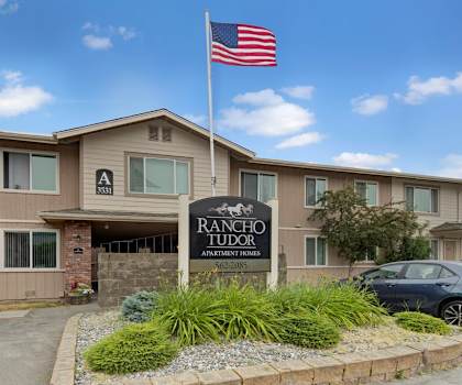 The exterior sign with trees and surrounded by greenery with the buildings and an American Flag in the background of Rancho Tudor Apartments. 