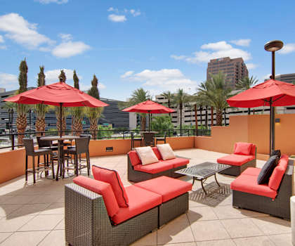 A patio with a red couch, soft red chairs. and dining tables and chairs with red umbrellas and palm trees in the background at Skyline Lofts Apartment Homes. 